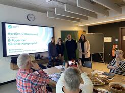 Petra,Theresa, der stellvertretende Chefredakteur und eine Mitarbeiterin der Berliner Morgenpost stehen rechts neben einem großen Fernseher und lächeln in die Kamera. Auf dem Fernseher ist die Startfolie der Präsentation zu sehen. Drei ältere Personen sitzen an einem Tisch mit kleinen Getränkeflaschen und Gebäck.