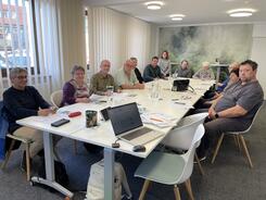 Die Teilnehmenden sitzen an einer langen Tischreihe und l&auml;cheln in die Kamera, Mobilreferentin Theresa sitzt hinter ihnen auf einem h&ouml;heren Hocker und l&auml;chelt ebenfalls.