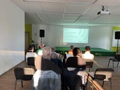 Die Teilnehmenden sitzen links in einem gr&ouml;&szlig;eren Saal, sind von hinten zu sehen und schauen auf eine Leinwand.