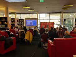 Die Teilnehmenden sitzen auf St&uuml;hlen und auf Sofas und sind von hinten zu sehen. Theresa referiert zu Online-Banking. Neben ihr ist die Leinwand mit Pr&auml;sentation.