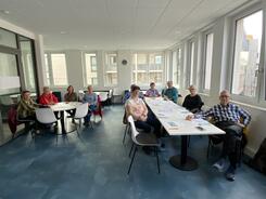 In einem Raum sitzen an einem langen Tisch und an einem runden Tisch insgesamt 11 Senior:innen und blicken nach vorne in die Kamera.