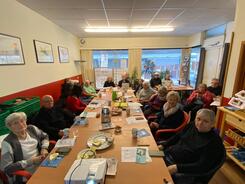 In einem recht kleinen Raum sitzen zwanzig Senior:innen an einem langen Tisch. Auf dem Tisch liegen gedruckte Bl&auml;tter und Getr&auml;nke. 