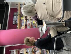 Mobilreferentin Theresa sitzt mit einer &auml;lteren Frau und dem Radiojournalisten an einem Tisch. Alle sind von hinten oder der Seite zu sehen. Im Hintergrund sind B&uuml;cherregale.