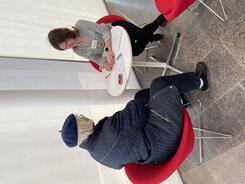 Theresa und eine &auml;ltere Besucherin sitzen auf roten St&uuml;hlen an einem wei&szlig;en runden Tisch. Theresa zeigt Dinge auf einem Zettel.