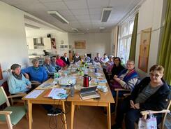 An einem langen Tisch sitzen ausschlie&szlig;lich &auml;ltere Frauen und blicken nach vorne in die Kamera. Auf dem Tisch liegen Getr&auml;nke und Materialien.