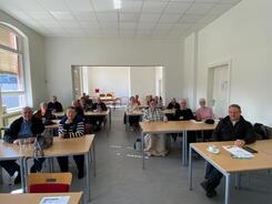 In einem Schulraum sitzen knapp 17 Senior:innen an Schultischen, auf denen Materialien liegen.