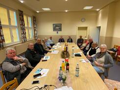 Im Dorfgemeinschaftshaus sitzen an zusammengestellten Tischen 14 Seniorinnen und Senioren und blicken nach vorne in die Kamera. Auf den Tischen liegen Unterlagen und Getr&auml;nke.