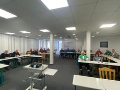 In einem gro&szlig;en Raum mit zwei Pfeilern in der Mitte sitzen Menschen an Tischen in U-Form. Im Vordergrund steht ein Beamer.