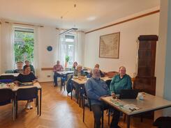 Mehrere Personen sitzen an Tischen verteilt im Raum und blicken nach Vorne.