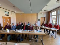 In einem Raum sitzen an f&uuml;nf Tischen insgesamt 14 Personen, die meisten davon Frauen. Sie blicken nach vorne in die Kamera. Auf den Tischen liegen Unterlagen und Kaffeetassen.