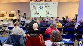 Die Teilnehmenden sitzen auf St&uuml;hlen im Computermuseum und sind von hinten zu sehen. Theresa steht zwischen Leinwand und alten Computern und l&auml;chelt in die Kamera.