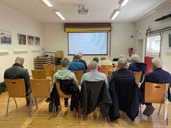 Die 10 Teilnehmenden und der Kooperationspartner sitzen in drei Stuhlreihen hintereinander, sind von hinten zu sehen und schauen auf die Leinwand mit der Startfolie der Pr&auml;sentation.