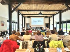 Die Teilnehmenden sitzen an Tischreihen in einem Fachwerkhaus mit Stelzen und schauen zu Theresa, die die Arbeit des Digitalen Engels vorstellt.