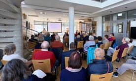 Die Teilnehmenden sitzen eng zusammen im Raum des Museums und sind von hinten zu sehen. Vorne steht Theresa links neben der Leinwand mit der PowerPoint-Folie zu schwacher und starker KI.