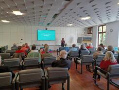 Theresa steht rechts neben dem Smartboard und stellt vor, was das Online-Banking ist. Die Teilnehmenden sitzen auf St&uuml;hlen in einem gr&ouml;&szlig;eren Rathaussaal und sind von hinten zu sehen.