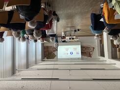 Theresa und die Mitarbeiterin des Landkreises l&auml;cheln in die Kamera. Die Teilnehmenden sitzen in Stuhlreihen und sind von hinten zu sehen.