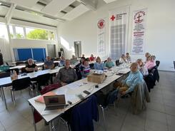 In einem Raum des DRK sitzen an zwei langen Tischreihen knapp unter 20 Senior:innen. Einige schauen in die Kamera.