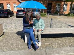 Mobilreferentin Petra (links) sitzt mit Seniorin Wilma auf einer Stra&szlig;enbank. Petra h&auml;lt einen blauen Schirm als Sonnenschutz &uuml;ber die beiden.