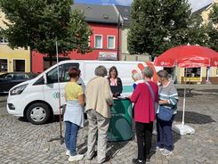 Mobilreferentin Theresa zeigt 5 &auml;lteren Frauen etwas auf dem Tablet. Sie stehen am Infostand auf einem Marktplatz. 