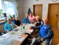 An einem Tisch sitzen sechs Menschen (vier Frauen links, zwei M&auml;nner rechts) und l&auml;cheln in die Kamera.