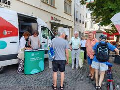 Mehrere Personen stehen auf einem Platz vor dem Infomobil, eine Dame l&auml;sst sich von Referent Johannes etwas auf ihrem Tablet zeigen.