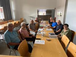 An einem langen Tisch sitzen acht Personen und gucken in die Kamera. Auf den Tischen liegen Unterlagen, digitale Ger&auml;te und Wasser.