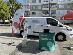 Das Infomobil inklusive Infostand stehen auf einem Marktplatz vor einem Mehrfamilienhaus.