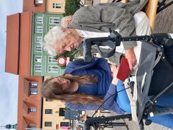 Petra (links) sitzt mit einer &auml;lteren Dame (rechts) auf einer Bierbank. Die Dame h&auml;lt ein Smartphone, auf das beide blicken. Vor den beiden steht ein Rollator.