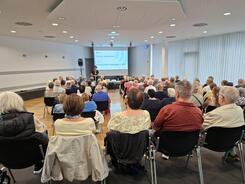 Zahlreiche Menschen sitzen auf St&uuml;hlen verteilt in einem gro&szlig;en Raum und blicken nach Vorne zu einer Leinwand.
