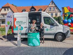 Eine Dame steht neben Johannes vor dem Infomobil und blickt auf eine Brosch&uuml;re, in der Johannes ihr etwas zeigt.