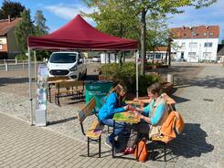 Petra sitzt auf einer Bierbank (links) mit zwei &auml;lteren Damen (rechts). Sie erkl&auml;rt einer Dame etwas auf dem Smartphone. Im Hintergrund steht ein Pavillon und das Info-Mobil.
