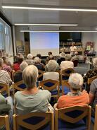 Zahlreiche Personen sitzen auf St&uuml;hlen im Raum und blicken nach Vorne auf eine Leinwand.