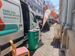 Das Infomobil steht auf einer Stra&szlig;e, vor dem Mobil sind ein Messetresen und St&uuml;hle. 