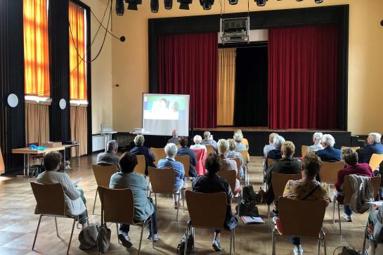 Senioren sitzen im Saal und hören einer Präsentation zu