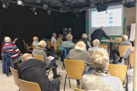 Senioren in Flensburg sitzen im Raum und schauen auf Leinwand