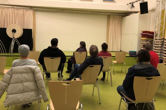 Teilnehmer sitzen im Saal und schauen auf Leinwand