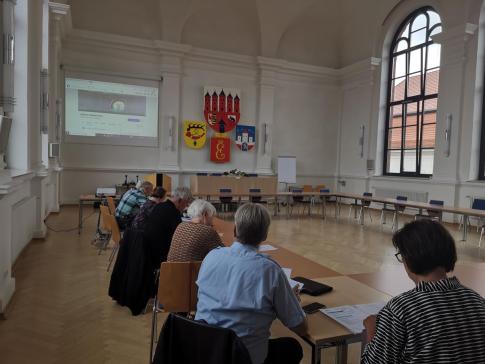 Personen sitzen an Tischen im Rathaus und blicken zur Leinwand