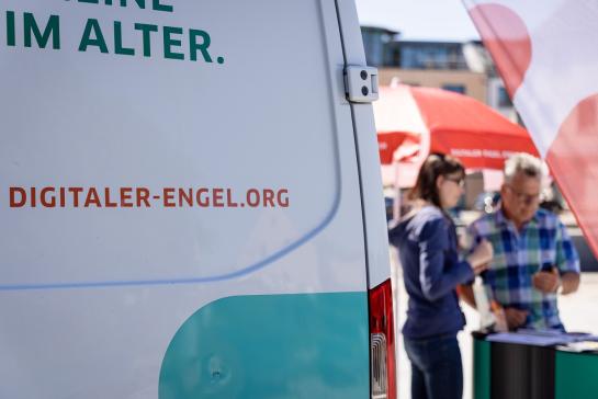 Im Vordergrund das Engel-Mobil mit der Aufschrift digitaler-engel.org. Im Hintergrund spricht Petra Rollfing mit einem Herr.