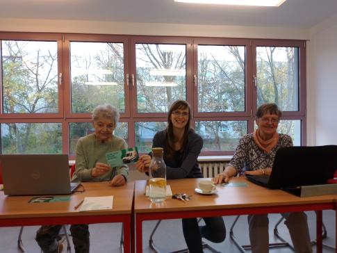 Die Referentin sitzt mit zwei Teilnehmerinnen an einer Tischreihe und lächeln in die Kamera. Vor beiden Teilnehmerinnen stehen Laptops. 