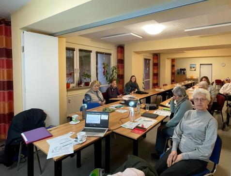 Sechs Senior:innen, Petra und Theresa sitzen an Tischen und lächeln in die Kamera.