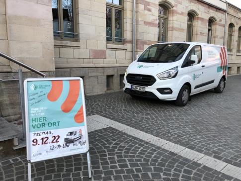 Das Engel-Mobil steht vor dem Bahnhofsgebäude. Im Vordergrund steht ein Aufsteller mit der Information, dass am 09.12.2022 hier der Digitale Engel zu Besuch ist.