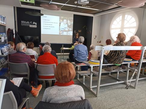 Senior:innen sitzen auf Stühlen und Bänken (von hinten fotografiert). Der Blick richtet sich nach vorne auf die Referentin vom Digitalen Engel mit einer Präsentation.