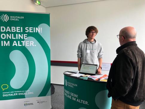 Mobilreferent Johannes ist im Gespräch mit einem Fachbesucher am Messestand des Digitalen Engel. Auf der linken Seite steht ein Roll Up mit der Aufschrift "Dabei sein! Online im Alter."
