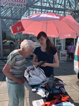 Petra zeigt einer Seniorin draußen am Infostand Dinge am Smartphone.