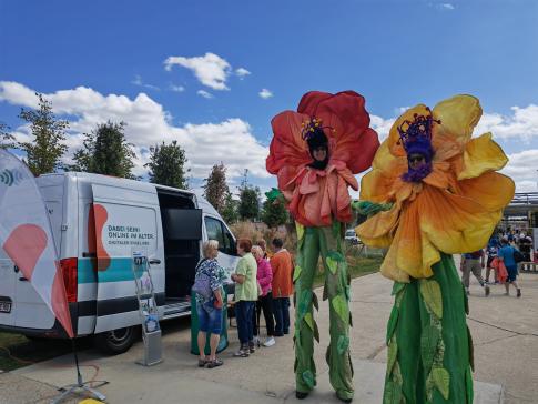 Zwei als übergroße Blumen verkleidete Maskottchen des Landesseniorentages stehen neben unserem Infomobil an dem sich einige Besucherinnen mit Johanna unterhalten 