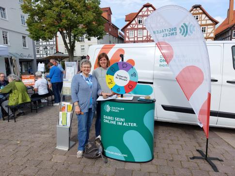 Theresa und eine Besucherin lächeln in die Kamera, links neben unserem Mobil sitzen einige Senior:innen an einem Tisch auf 2 Partybänken.