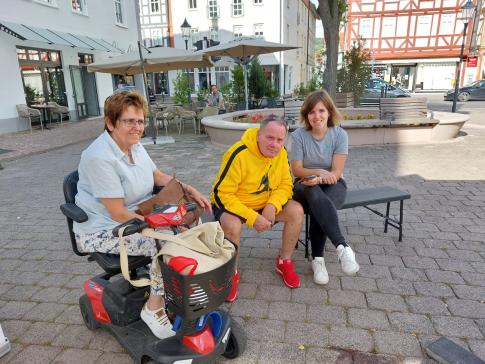 Theresa und ein Besucher sitzen auf der Bank, links neben ihnen eine Seniorin in einem elektrischen Rollstuhl.