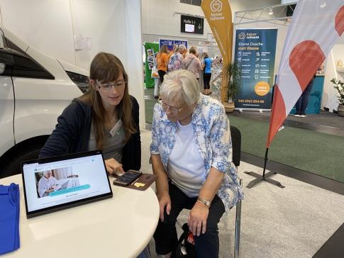 Petra sitzt mit Seniorin Christa an einem Tisch und zeigt etwas auf dem Smartphone. Neben den beiden steht auf dem Tisch ein Tablet mit der Internetseite www.digitalen-engel.org.