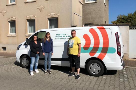 Petra steht mit Frau Allenfort von der Stiftung Finneck und einem Teilnehmer vor dem Info-Mobil. Sie posieren in die Kamera.