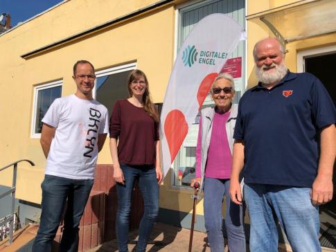 Quartiersmanager Stephan Reich, Referentin Petra Rollfing, eine Seniorin sowie Peter Brinkhus stehen vor dem MGH und schauen in die Kamera. In der Mitte steht die Beachflag vom Digitalen Engel.